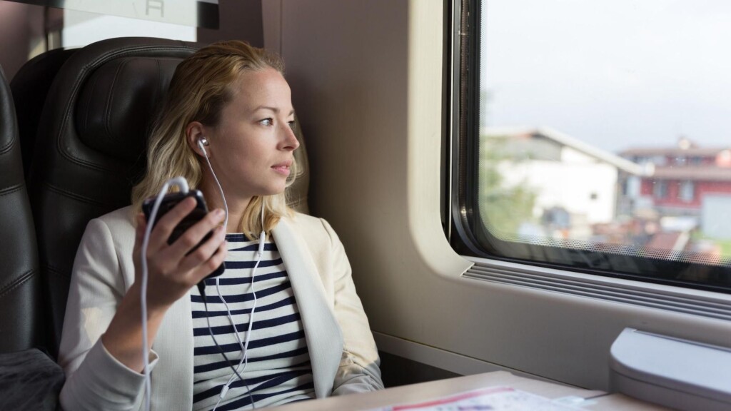 Eine aufmerksame Geschäftsfrau, die durch das Fenster schaut und beim Podcast auf dem Handy mit Kopfhörer zuhört, während sie mit dem Zug in der Business Class sitzt. (1120723976) - shutterstock.com